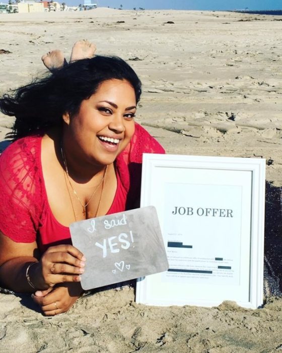 Benita Abraham acostada en la playa con una foto enmarcada que dice OFERTA DE EMPLEO y un letrero que dice DIJE QUE SÍ